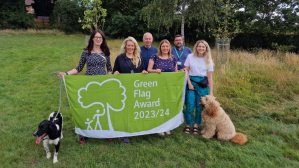 Six people in a grassy outdoor orchard holding a big flag with Green Flag Award 2023/4 written on it flanked by a collie dog and a fuzzy sandy dog